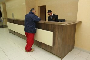 A reception desk with a man in a blue jacket and red pants standing at it, and another man in a black suit seated behind the desk.