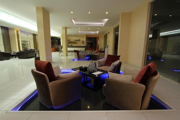 A modern hotel lobby with a central seating area featuring sofas and a coffee table, a kitchen in the background, and a glass partition wall.
