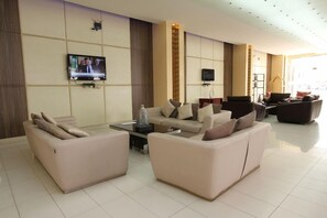 A modern hotel lobby with a seating area, two flat-screen TVs mounted on the wall, and a large window providing natural light.