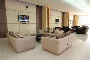 A modern hotel lobby with a seating area, two flat-screen TVs mounted on the wall, and a large window providing natural light.