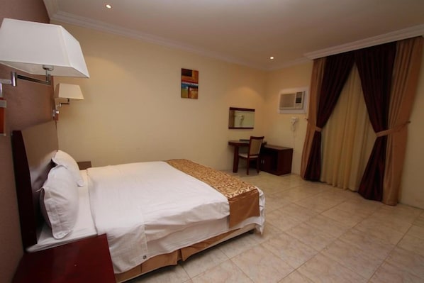 A hotel room with a bed, a desk, a chair, a lamp, and a window with curtains. - Raoum Inn Serviced Apartments Khafji (Al Khafji)