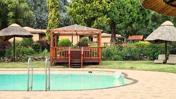 Ein Schwimmbecken mit Holzterrasse und einem Pavillon, umgeben von Bäumen und Strohdächern.