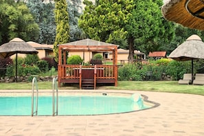 A swimming pool with a wooden deck and a gazebo, surrounded by trees and thatched umbrellas.