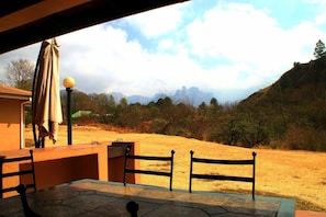 An outdoor seating area with a table and chairs, a lamp post, and a view of a hilly landscape with trees and a distant mountain range.