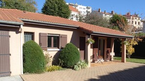 A two-story building with a tiled roof, a front porch with a bench, and a small garden area with shrubs. - Le Clos Gentiane [vr] (Saint-Etienne)