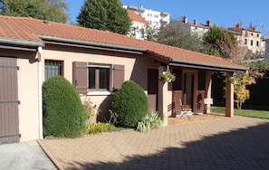 A two-story building with a tiled roof, a front porch with a bench, and a small garden area with shrubs. - Le Clos Gentiane [vr] (Saint-Etienne)