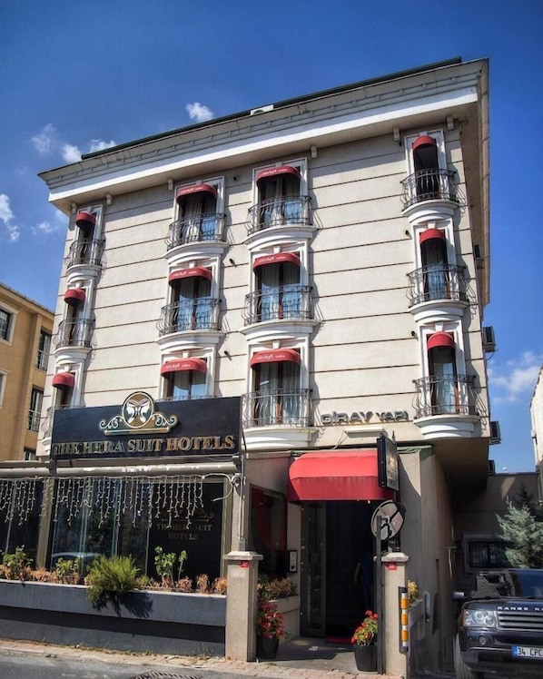 Un hotel de varios pisos con balcones, un toldo rojo y un cartel que dice 'The Hera Suit Hotels'.