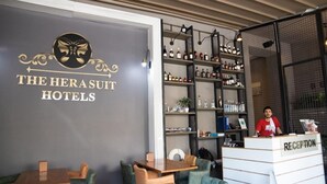 Ein Empfangsbereich mit einer Rezeption, an der 'RECEPTION' steht, einer Wand mit dem Logo und dem Namen 'THE HERASUIT HOTELS' sowie Regalen mit verschiedenen Flaschen.