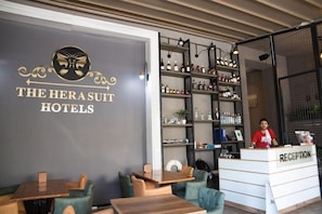 Un'area di accoglienza con una reception identificata da una targa 'RECEPTION', una parete con il logo e la scritta 'THE HERASUIT HOTELS' e mensole con diversi flaconi.
