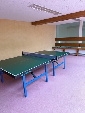 A ping pong table with blue legs and a green playing surface, positioned in a room with a wooden bench and a window in the background. - Schluchsee Dream in the Black Forest (Schlochsee)