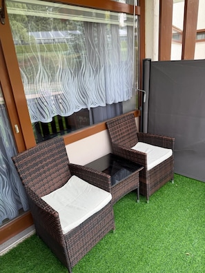 Ein Zimmer mit zwei Korbstühlen und einem kleinen Tisch, einem Fenster mit Spitzenvorhang und einem grünen, künstlichen Rasenbelag.