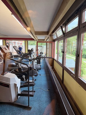 Ein Fitnessraum mit Cardiogeräten, einem blauen Teppich und großen Fenstern mit Blick auf Grünflächen.