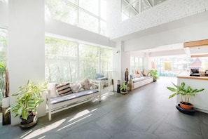 A spacious living area with a high ceiling, large windows, and a seating area with a sofa and a bench. - Capital O 631 Blooming Hill Boutique Suites (Pattaya)