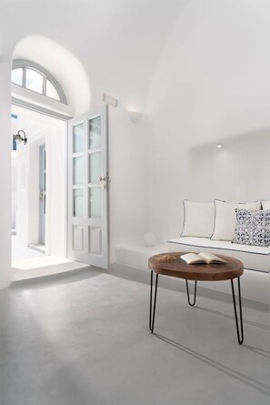 A modern interior with a white sofa, a round wooden table, and a white door leading to another room. - Oia Spirit Suites Adults Only (Santorini)