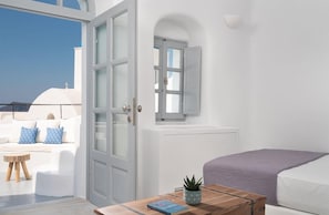A room with a bed, a small table with a book and a plant, a window with a view of the sky, and a door leading to an outdoor area with a white sofa and cushions. - Oia Spirit Suites Adults Only (Santorini)