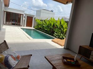 A poolside area with a lounge chair, a table with a tray of items, and a view of a building with a sliding door. - Rumah Melissa Modern Boho 3BR Villa in Sanur (Denpasar selatan)