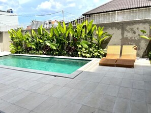 Piscina com deck ladrilhado, rodeada por plantas verdes exuberantes e duas cadeiras de vime.
