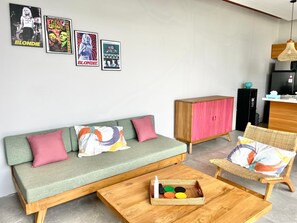 A modern living room with a green sofa, a wooden coffee table, and a wicker chair. There are framed posters on the wall and a cabinet with a pink door. - Rumah Melissa Modern Boho 3BR Villa in Sanur (Denpasar selatan)