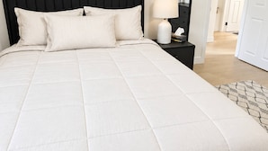 Una cama bien hecha con cabecera negra, una mesita de noche con lámpara y una obra enmarcada en la pared.