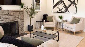 A modern living room with a fireplace, a flat-screen TV, a coffee table, and two armchairs.