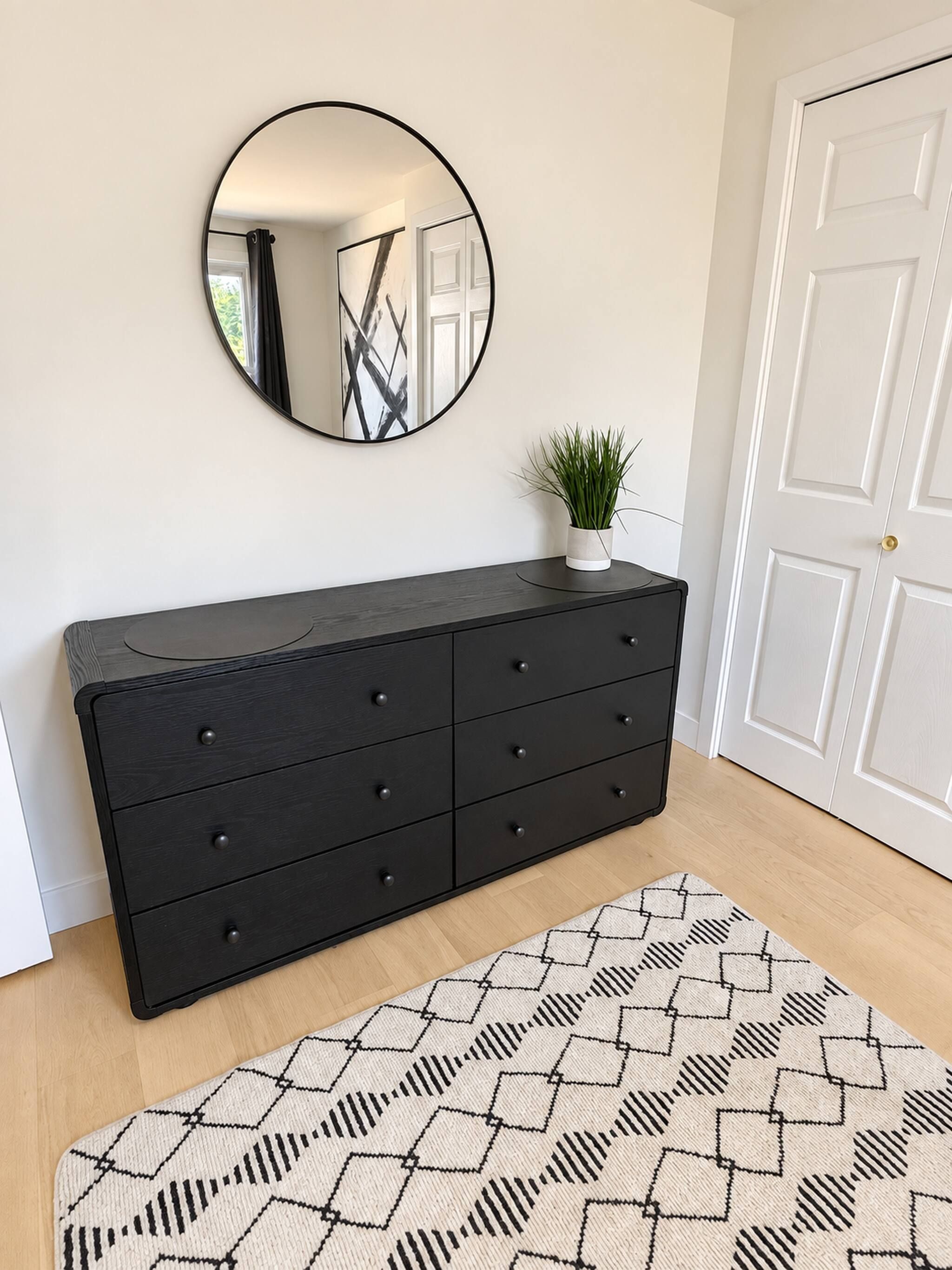Una habitación con un aparador de madera oscura, un espejo redondo, una planta en maceta y una alfombra estampada.