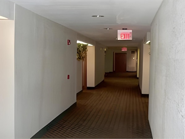 A hotel hallway with a carpeted floor, white walls, and an exit sign. - Scottish Inns (Houston)