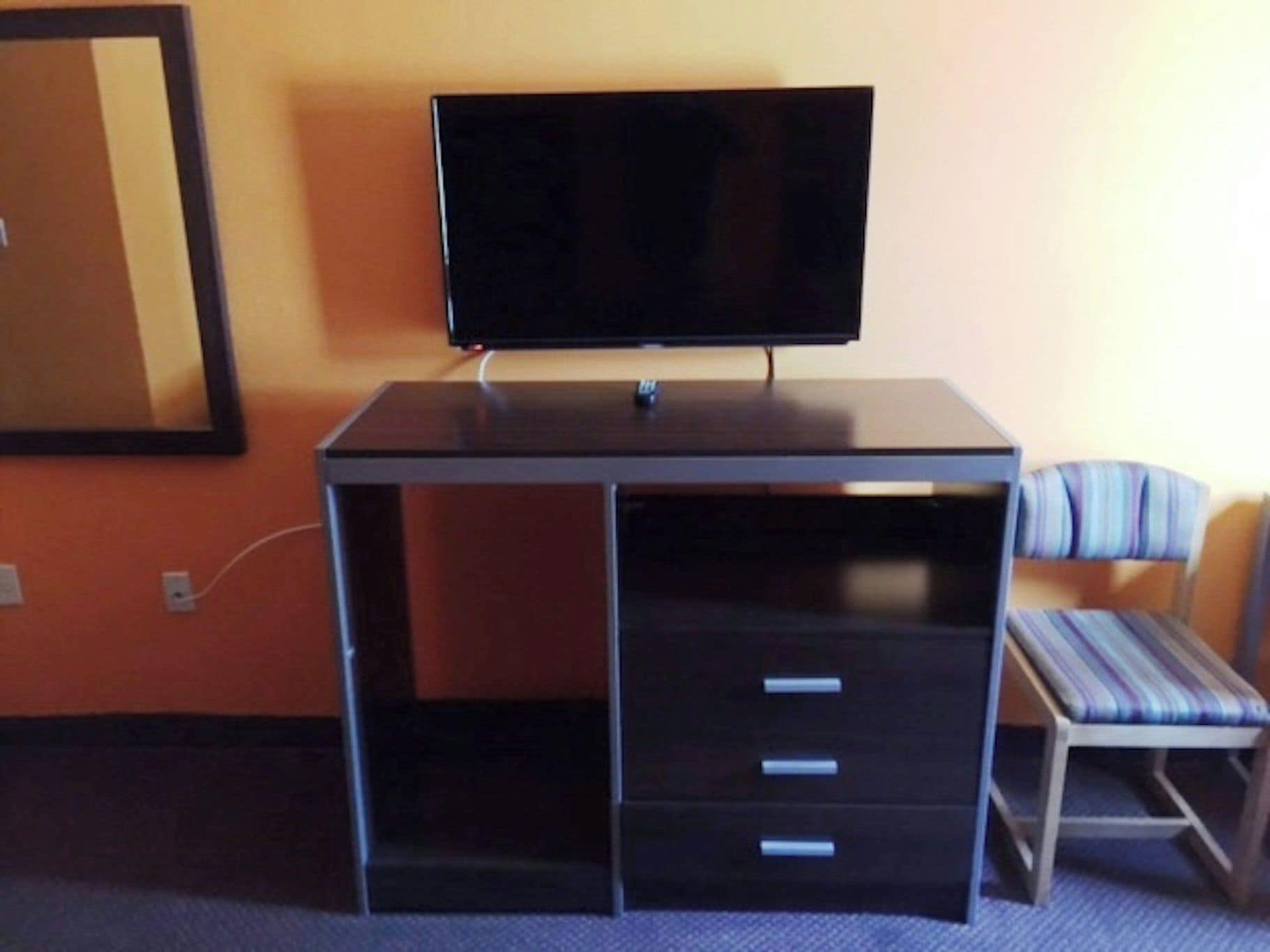 A room with a desk, a television on top, a chair with a striped cushion, and a mirror on the wall.