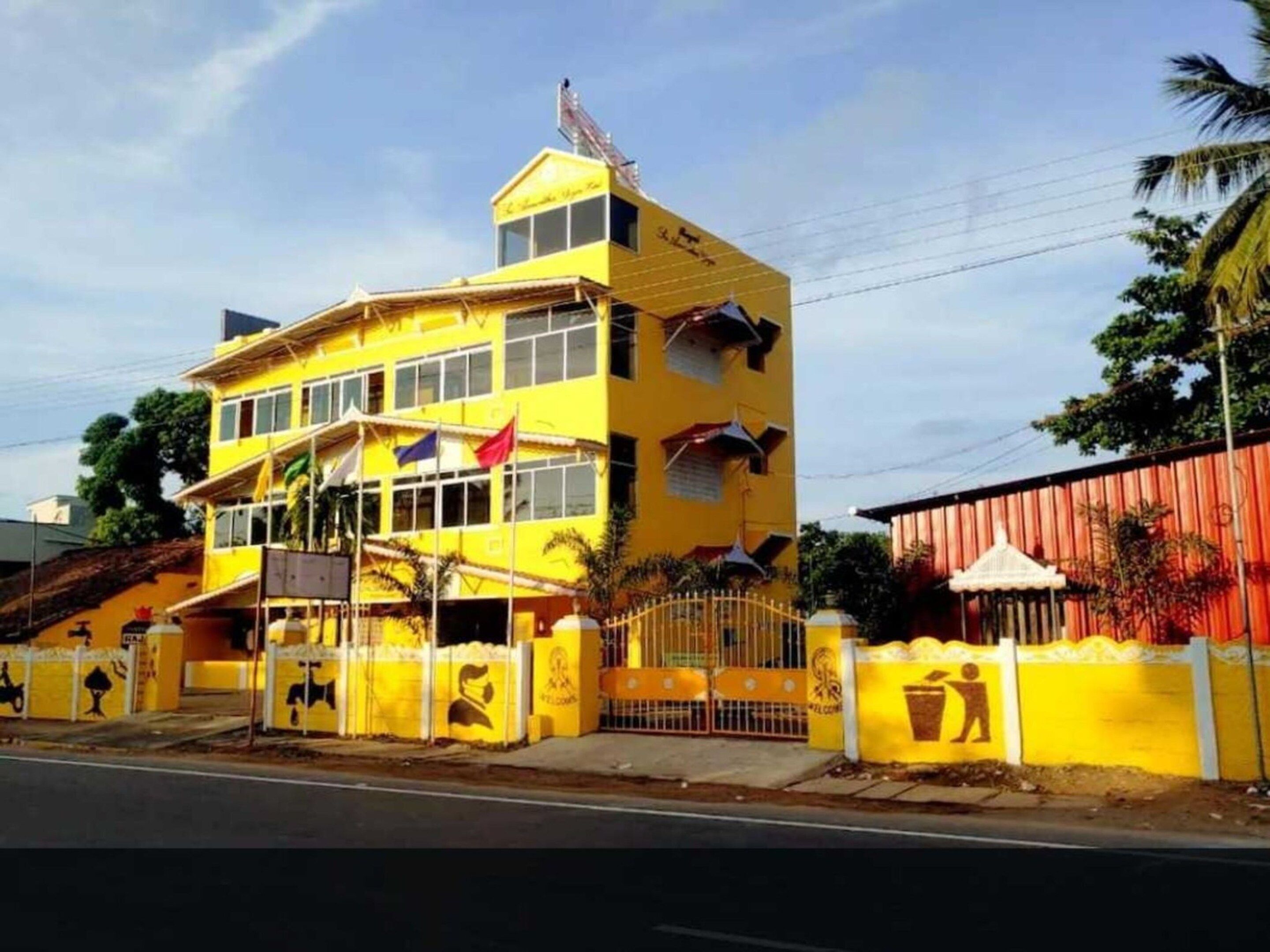 A yellow multi-story building with a flag on top, a gated entrance, and a fenced area with waste bins.