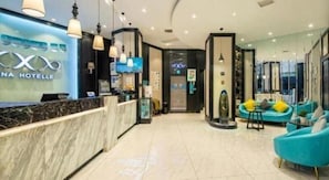 Zona de recepció d'hotel amb mostrador de marbre, taulell de recepció i espai d'estar amb cadires blaves.