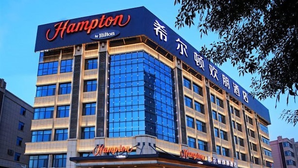 Un hotel con un llamativo letrero de 'Hampton by Hilton', varios pisos y una entrada amplia con un cartel luminoso.