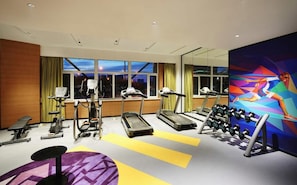 A gym with cardio machines, dumbbells, and a large window with a city view.