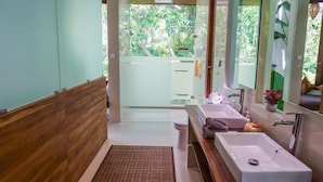 Ein Badezimmer mit Holzboden, einem großen Spiegel, zwei Waschbecken, einer Toilette und Blick ins Grüne.