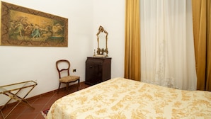 Ein Schlafzimmer mit einem Bett, einem Sessel, einem Schminktisch und einem Spiegel. An der Wand hängt ein Gemälde.