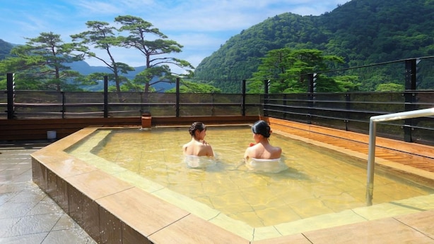 Ein Thermalbad mit zwei Personen im Wasser, umgeben von einer Holzterrasse und einem Metallgeländer, mit einer Berglandschaft im Hintergrund.