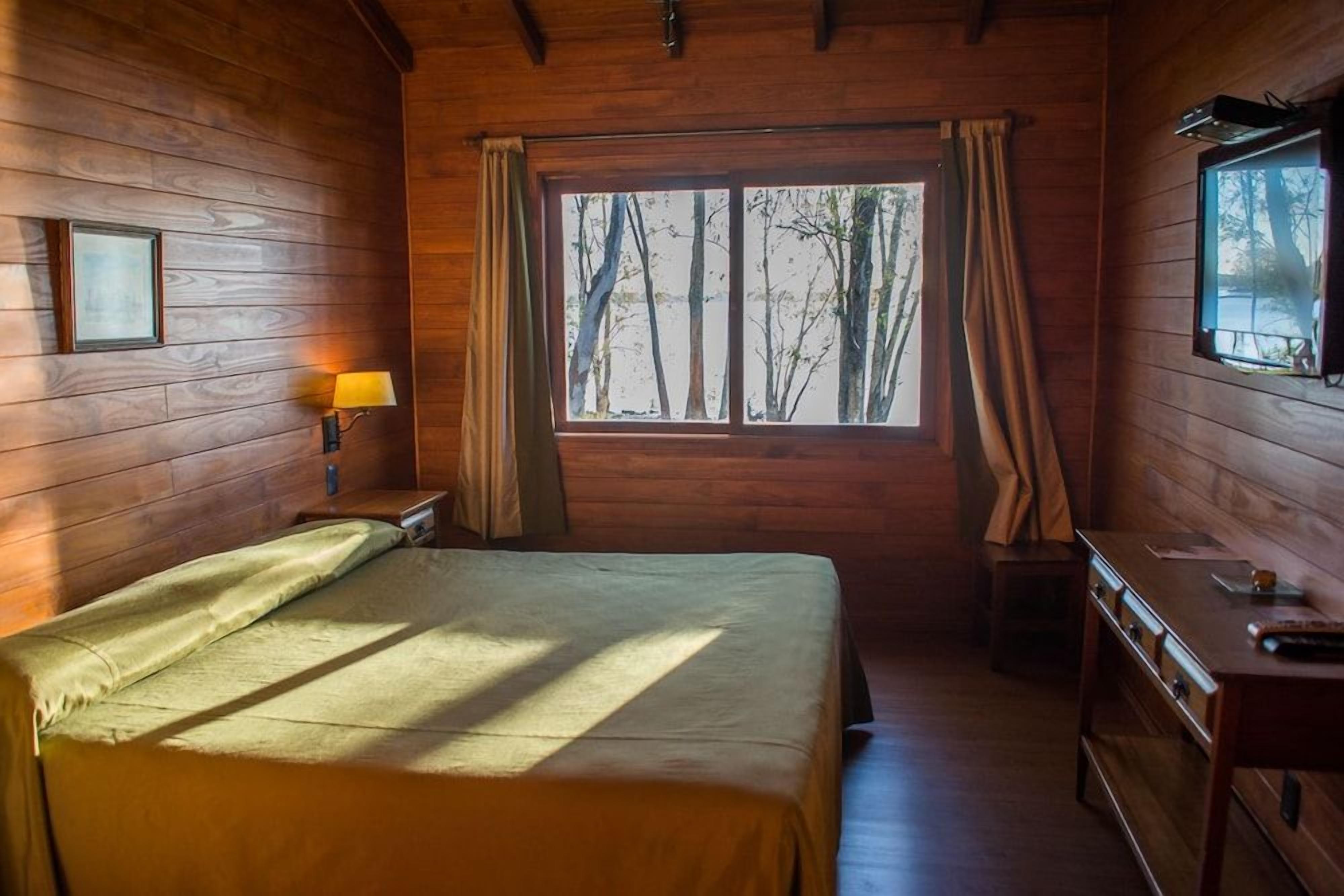 A wooden cabin room with a bed, a desk, and a window with curtains.
