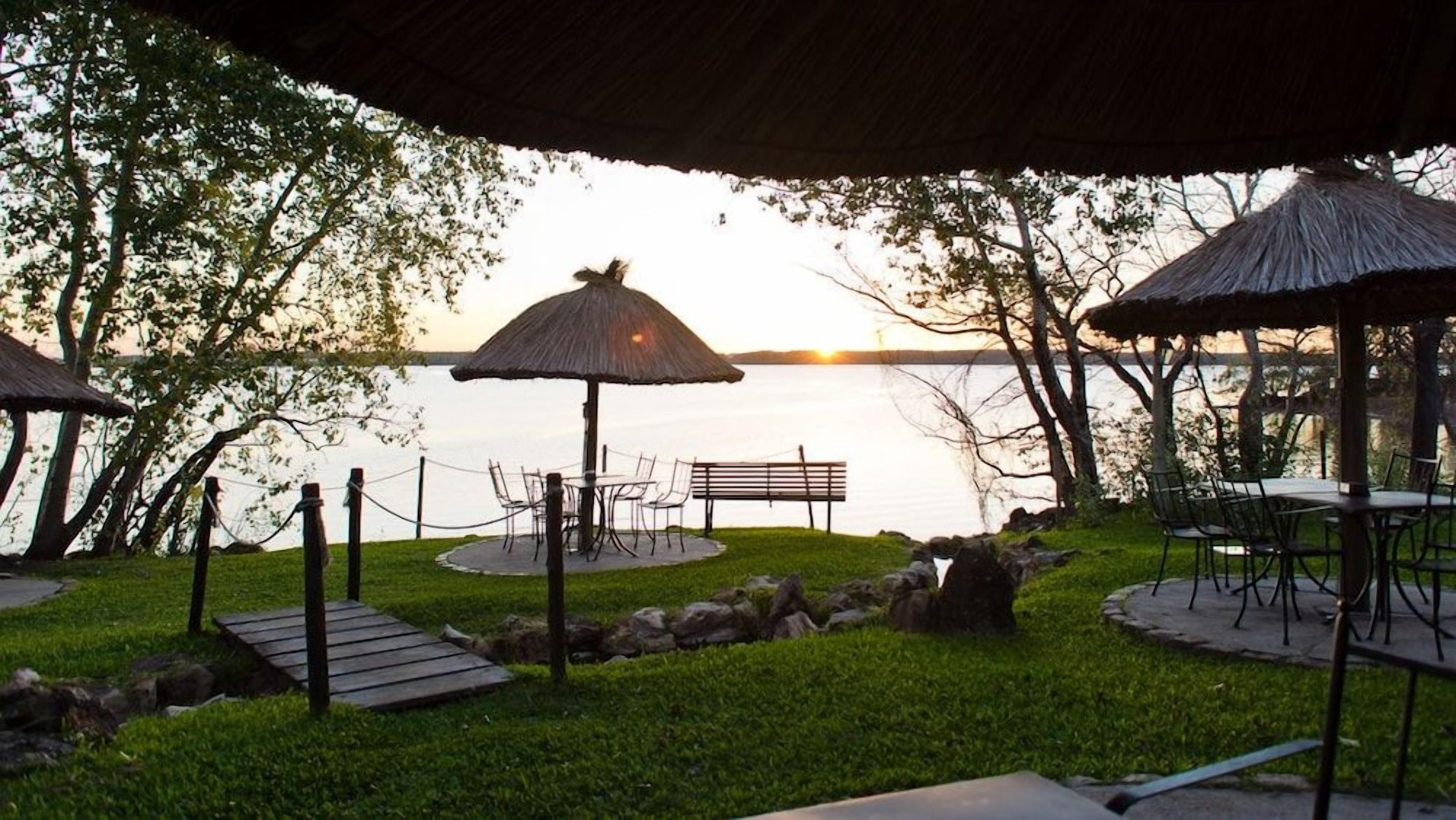 An outdoor seating area with tables and chairs, surrounded by a rope barrier, near a body of water with a sunset in the background.