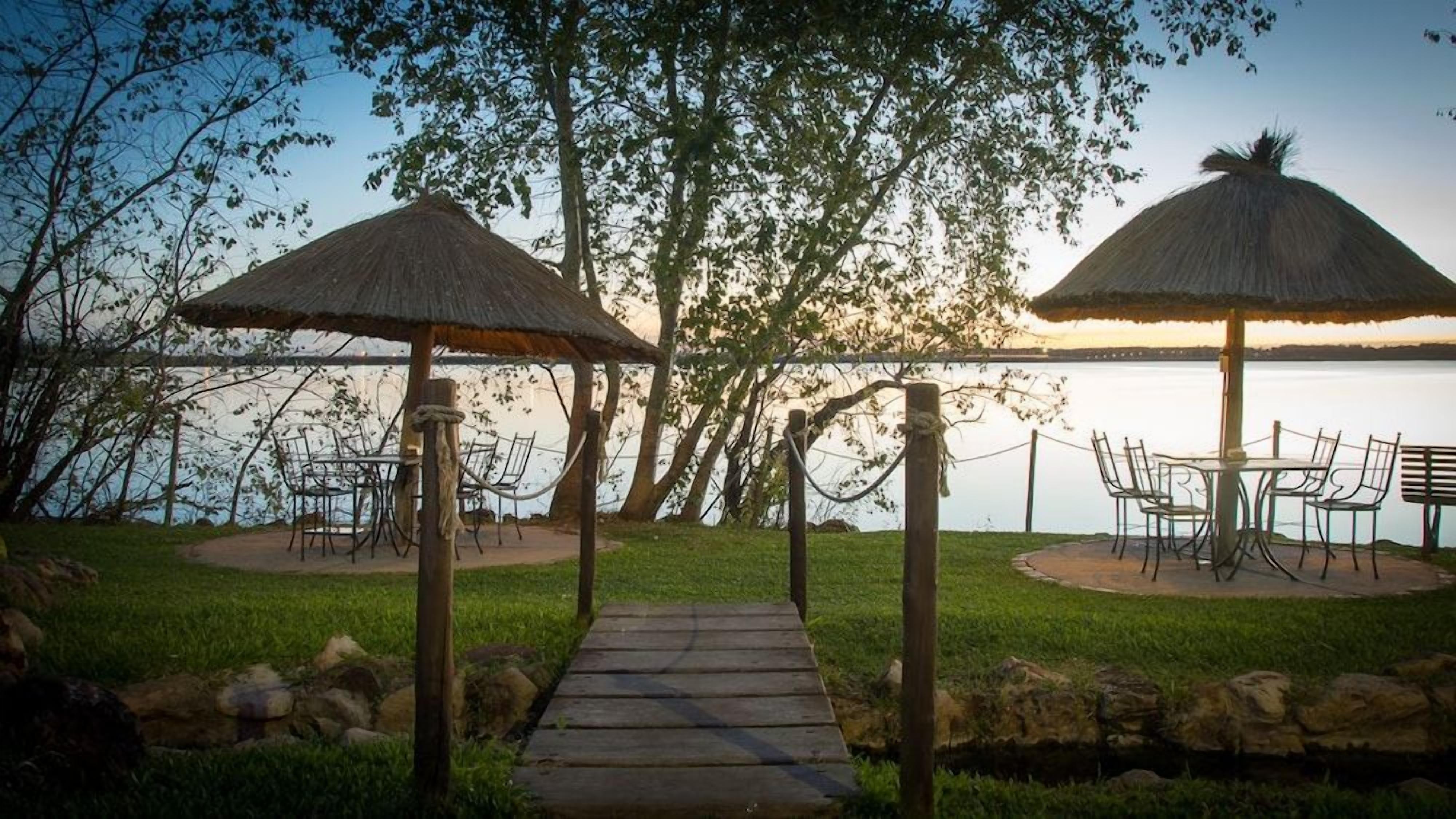 A wooden pathway leading to two outdoor seating areas with thatched umbrellas, overlooking a body of water.