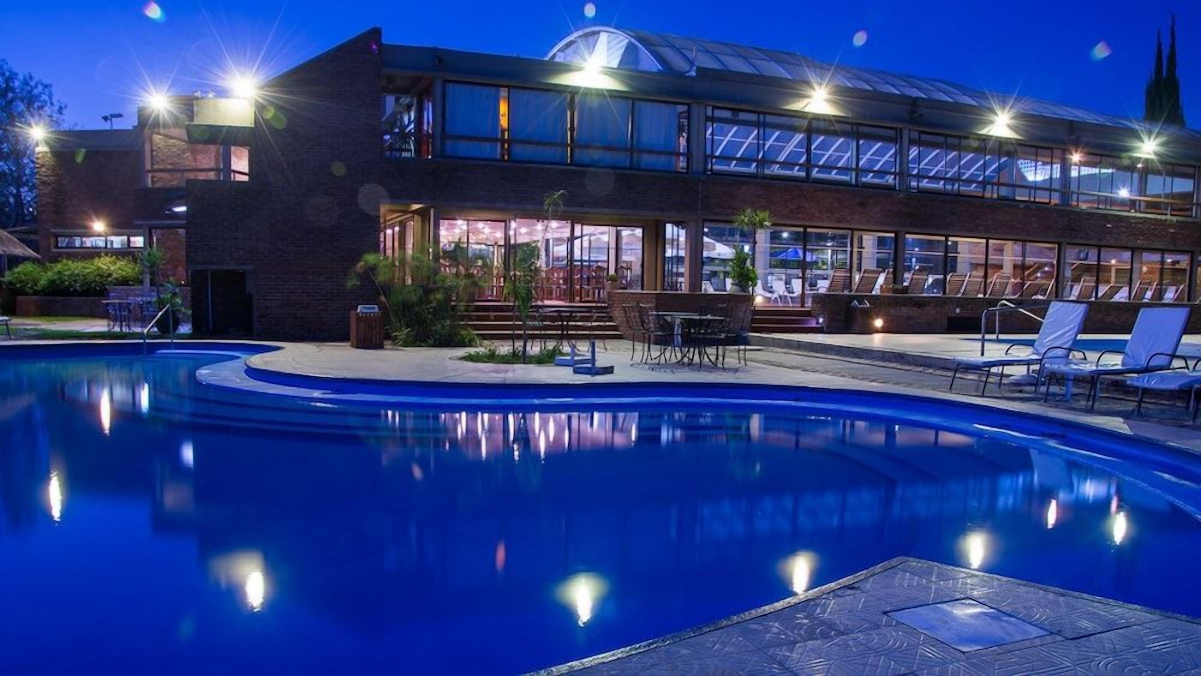 A swimming pool with lounge chairs and tables, a covered outdoor seating area, and a building with large windows and a glass roof.