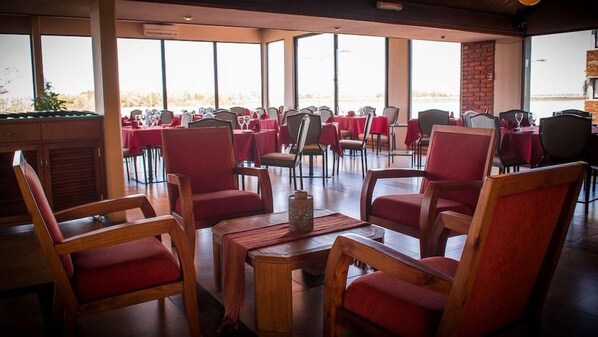 A dining area with red upholstered chairs and tables, large windows, and a brick wall.