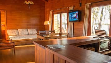 A wooden cabin interior with a sofa, dining table, chairs, a television, and a built-in oven.