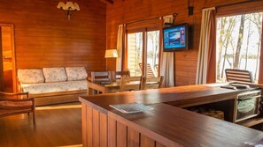 Interior de una cabaña de madera con sofá, mesa de comedor, sillas, televisor y horno empotrado.
