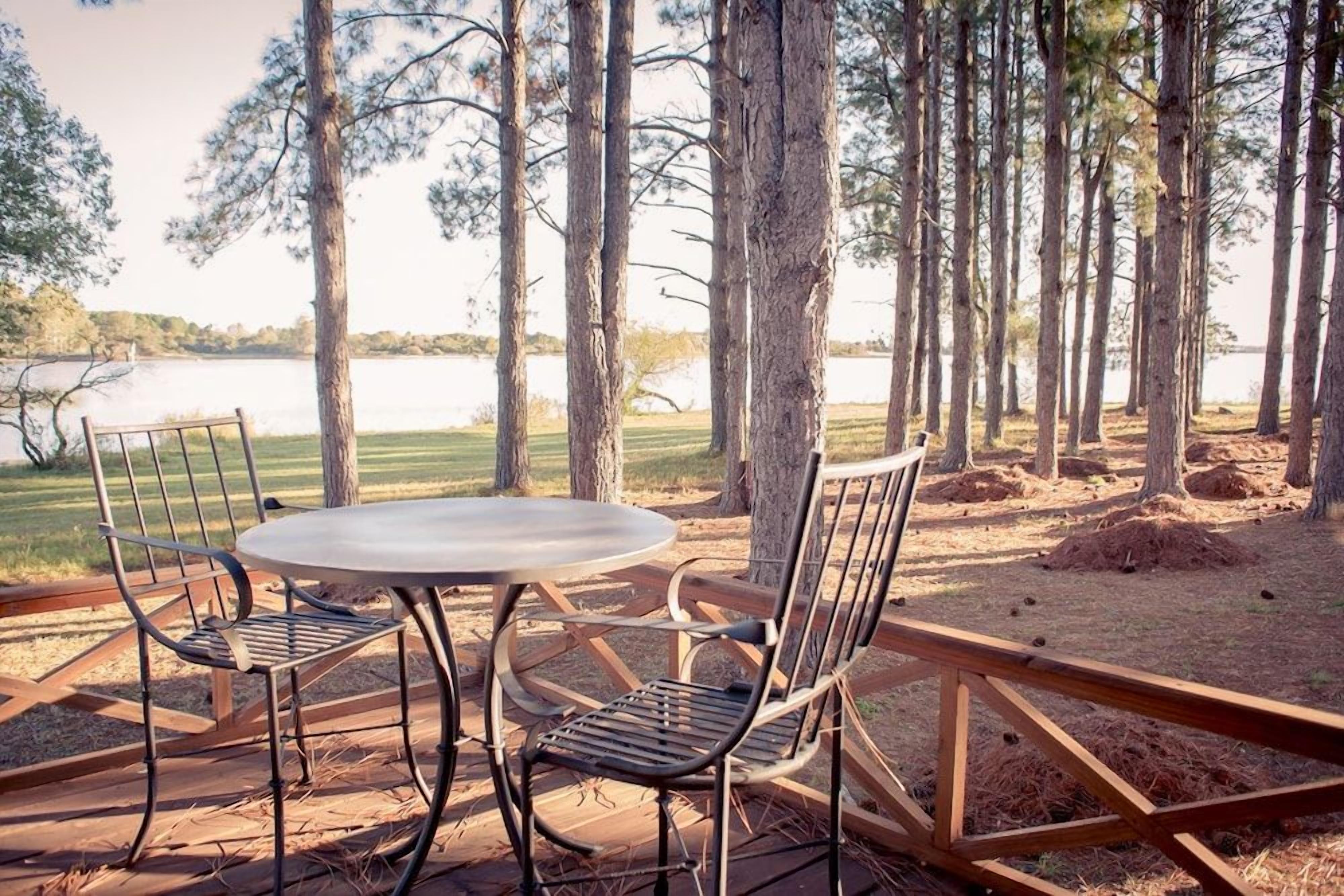 An outdoor seating area with a round table and metal chairs, surrounded by trees and a view of a lake.