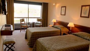 A hotel room with two beds, a desk with a chair, a small table, and a window with curtains.