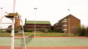 Una cancha de tenis con una red, rodeada por una valla, y un edificio con varias ventanas y un techo verde al fondo.