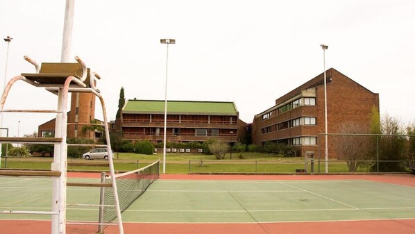 Una cancha de tenis con una red, rodeada por una valla, y un edificio con varias ventanas y un techo verde al fondo.