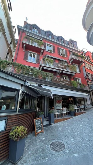 Um edifício vermelho com restaurante no andar térreo, placa com 'Hotel La Rouvière' e varanda com plantas.
