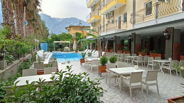 Zona de piscina exterior con sillones y mesas, rodeada de vegetación y edificios.