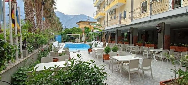 Zona de piscina exterior con sillones y mesas, rodeada de vegetación y edificios.