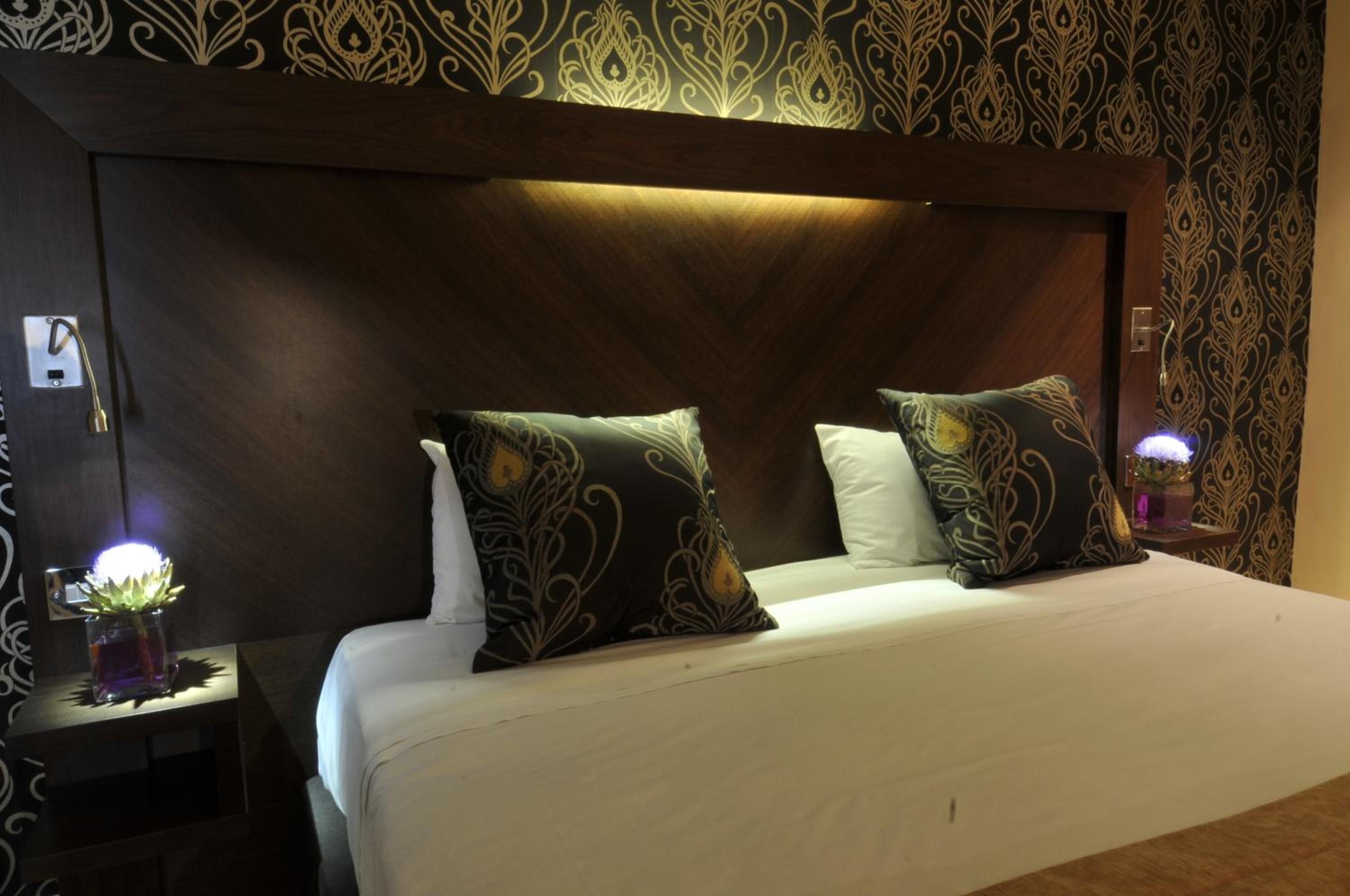 A hotel room with a large bed, two decorative pillows, a nightstand with a lamp, and a wall with an ornate pattern.