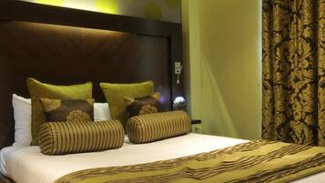 A hotel room with a large bed, multiple pillows, a headboard, and a patterned curtain.